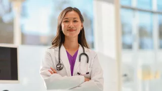 A female doctor stands in a hospital, with her back to a computer screen, with natural light streaming in from windows behind her.